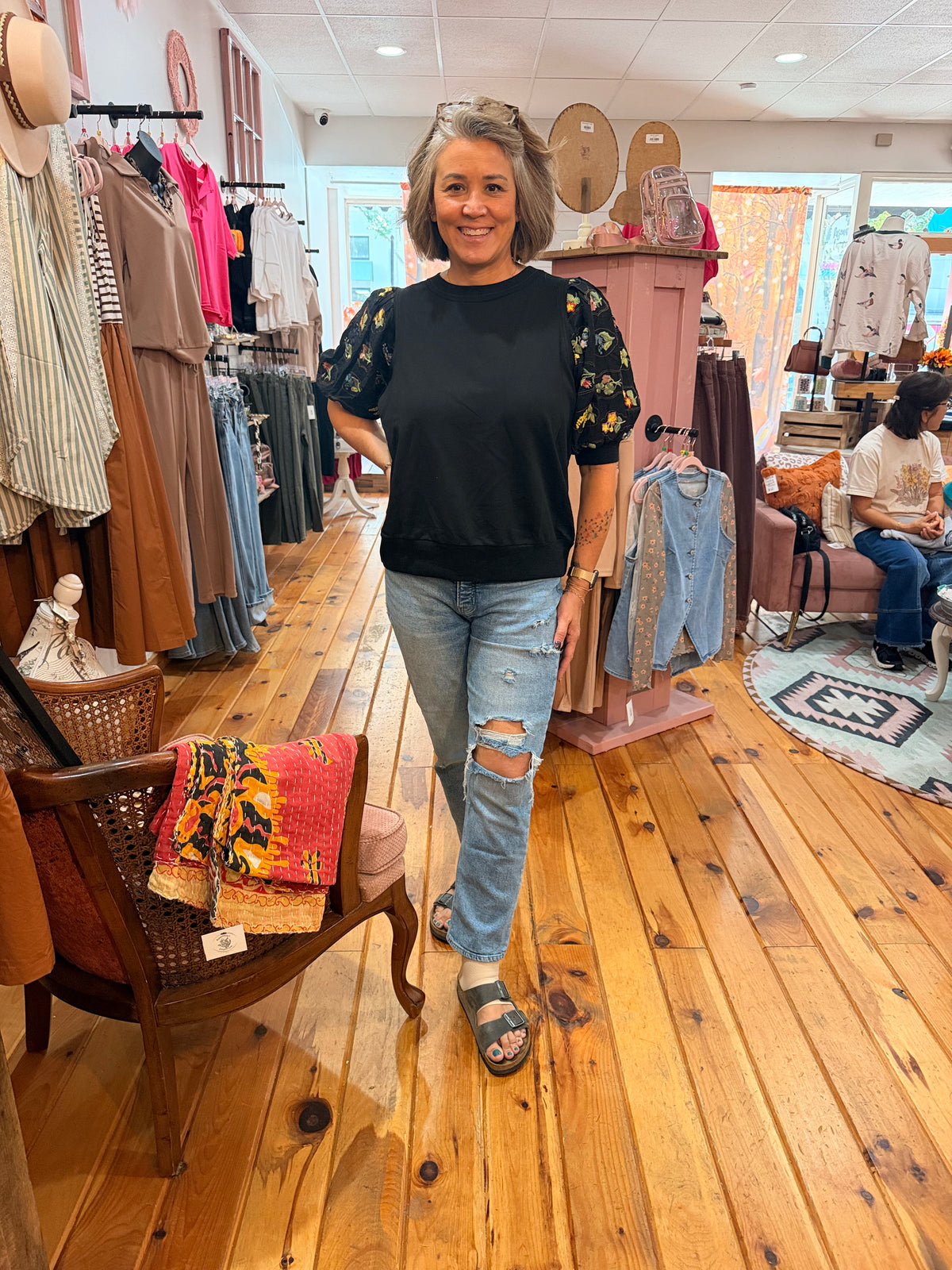 A woman wearing a black crewneck top with floral puff sleeves and light-wash distressed jeans stands in a boutique with wooden floors and clothing racks.